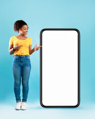 Cheerful black woman standing next to huge smartphone, pointing at empty screen, advertising new mobile application or website. Blue studio background, cellphone display mockup