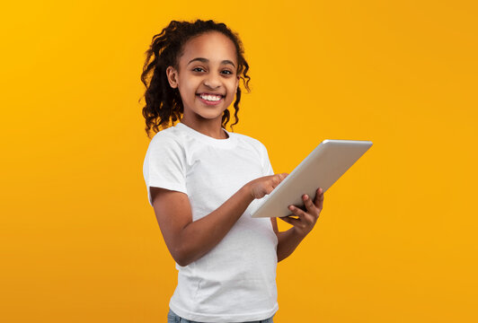 Digital Natives, App For Kids. Beautiful smiling African American teenage girl holding and using digital tablet over orange studio background, looking and posing at camera, banner with copy space - Powered by Adobe