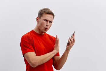 Young man wearing red t-shirt, looking confused while using smartphone, isolated on plain light gray background, studio portrait, expressing surprise or disbelief. People lifestyle concept