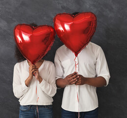 Love and Valentines day. African-american couple hiding faces behind red heart shaped balloons,...