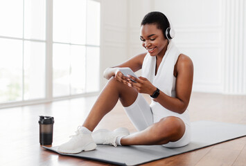 Obraz premium Happy young African woman with towel on shoulders and wireless headset sitting on fitness mat in living room, listening to music while resting, free copy space. Sports music mobile application concept