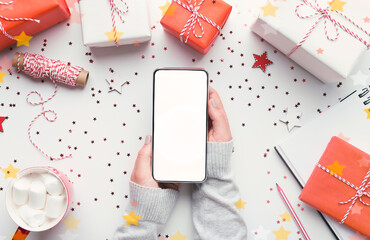 Online Christmas shopping. Modern cellphone with white blank screen in woman hands on background with gifts, copy space