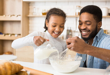 Cute African American Girl Pouring Milk Into Bowl At Kitchen, dad mixing the dough, copy space