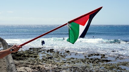 Hand holding a Palestine flag on a beach in the Canary Islands, expressing support for a humanitarian aid flotilla sailing to Gaza, with surfers enjoying the ocean waves in the background