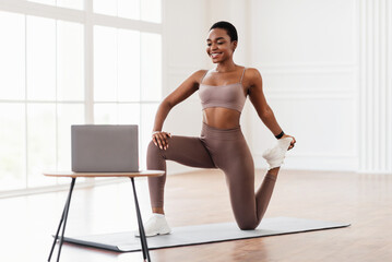 Fototapeta premium Your Personal Trainer. Smiling female fitness instructor showing legs quad stretching exercises while streaming, broadcasting video lesson using laptop, looking at screen. Sport, online gym concept