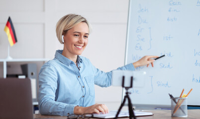 Online languages school. Millennial female tutor giving German lesson on internet, using smartphone, pointing at blackboard, explaining grammar to remote students. Education during covid-19 epidemic