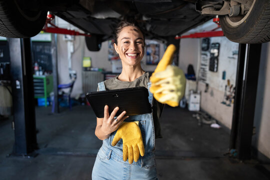 A joyful mechanic shares a thumbs-up while engaging with her tablet under a vehicle, highlighting her enthusiastic work