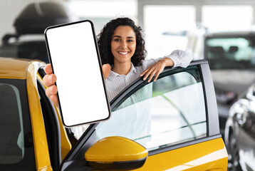Positive millennial middle eastern woman standing by brand new yellow car, showing smartphone with...