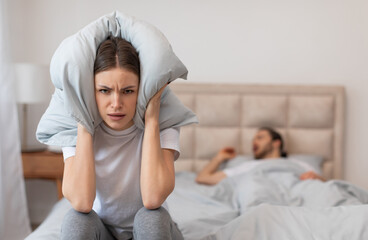 Exasperated woman sitting in bed holding pillow over her ears to block out the sound of man snoring loudly in the background, focus on angry lady