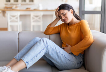 Sad young indian woman suffering from PMS and menstruation pain at home, upset eatsern female having stomach ache, acute abdominal pain and headache, sitting on couch in living room, copy space
