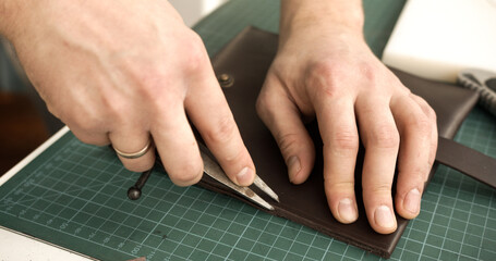 Leather artisan using divider tool to mark stitching line on brown leather wallet closeup