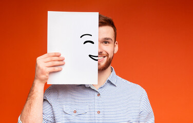Young smiling guy supplement half of his face with painting smile on white sheet of paper, panorama, copy space, orange background