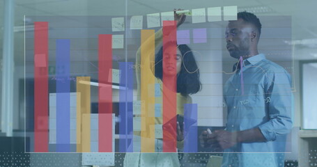 Collaborating colleagues adjusting notes on glass board at office, with smartphone, digital charts