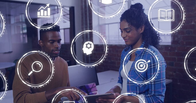 Gesturing male and female coworkers examining tablet near monitors at office, with floating icons