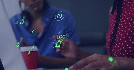 Collaborating team in formal wear analyzing data on laptop in office, environmental icons, red cup