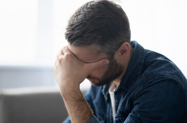 Upset middle-aged bearded man having headache, sitting on couch at home and touching his forehead,...
