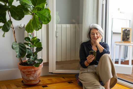Senior woman relaxing at home using smartphone by fiddle leaf fig plant