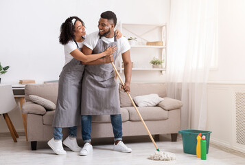 Happy black couple doing household chores together, mopping floor and hugging, spring-cleaning...
