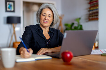 Fototapeta premium Senior woman working from home office taking notes using laptop computer