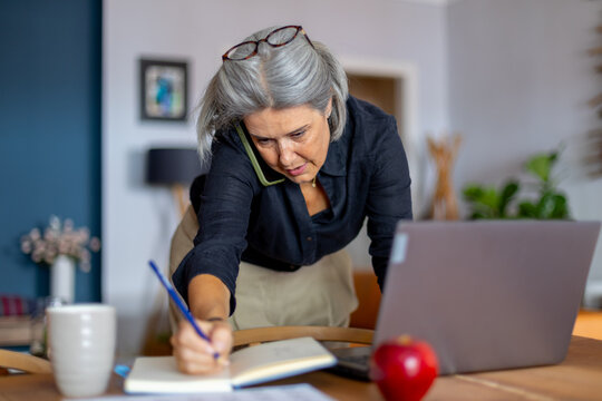 Focused senior woman managing business from home office, taking notes while on phone call