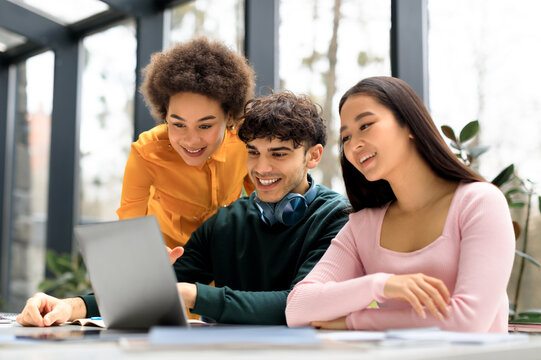 Happy international students studying in coworking space, enjoying the results of their project, looking at laptop screen and smiling, sitting together indoors