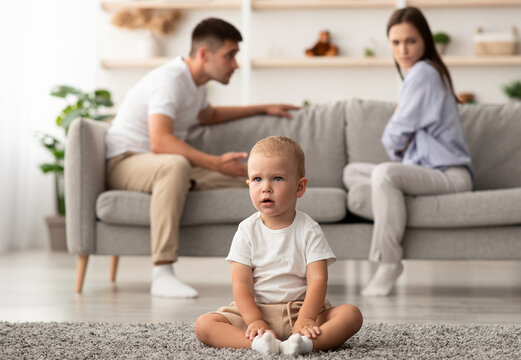 Domestic Quarrels Concept. Cute little infant baby boy sitting alone on floor separately from arguing parents at home, young family suffering relationship crisis, thinking divorse, selective focus