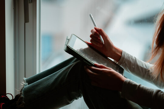 Asian woman freelancer sitting by the window, remote working on digital tablet in apartment room. University student online study, E-learning at home. Wireless technology, gadget device for working.