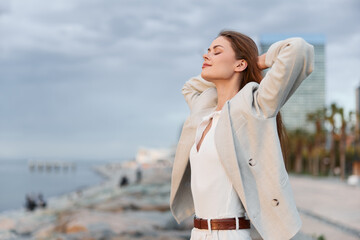 woman enjoying fresh air outdoors near waterfront, casual beige jacket and white outfit, serene...