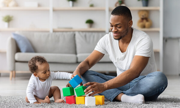 Happy Young Black Father Playing With His Little Infant Baby Son At Home, African American Dad And His Cute Toddler Male Child Stacking Building Blocks While Sitting Together On Floor In Living Room