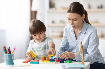 Fototapeta premium Preparing to school classes. Cute little boy preschooler playing developing wooden games with professional woman psychologist at kindergarten, free space