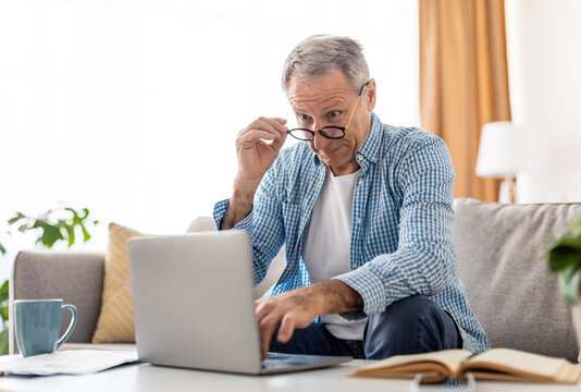 Omg, Wow. Portrait of surprised mature man sitting at desk on couch using laptop, taking off glasses looking at pc screen in amazement. Emotional male reading breaking news online in home office