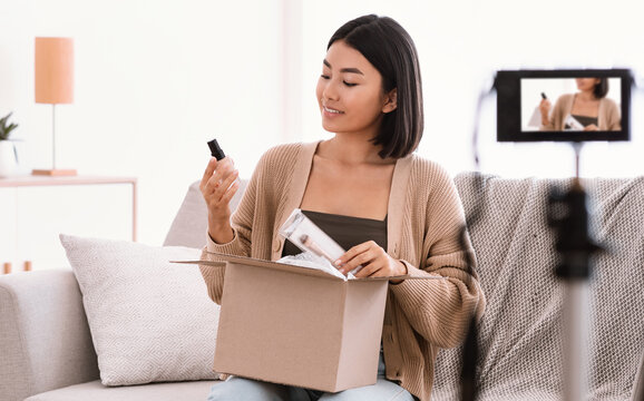 Product Review. Satisfied asian young woman vlogger holding foundation and brush, unpacking cardboard beauty box with cosmetics products, recording video for her social media blog on camera at home