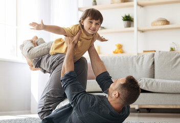 Happy young caucasian father lying on floor and carrying his little son on hands, kid playing plane...
