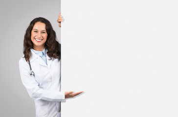 Cheerful female doctor with stethoscope peeking out from behind blank white board, presenting space...