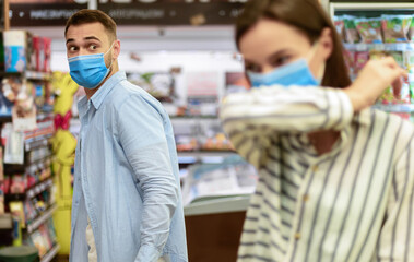 Obraz premium Sick Woman Coughing Or Sneezing In Elbow Wearing Mask Doing Grocery Shopping In Supermarket, Buying Food, Scared Guy Looking At Her. Coronavirus Covid-19 Pandemic Outbreak, Social Distancing