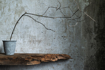 Minimal branch in metal pot on rustic wood shelf against textured wall, capturing wabi sabi beauty in natural simplicity.