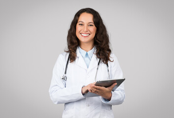 Positive woman in medical workwear doctor using digital tablet isolated on grey background, chatting with patient. Modern technologies in healthcare