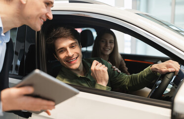 Young Caucasian couple clarifying car purchase details with salesman, looking at tablet screen in...