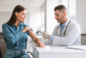 Inoculation time. Doctor man wearing gloves and making vaccination shot to young female patient, inserting syringe to her arm, woman getting vaccine shot against viruses in modern clinic