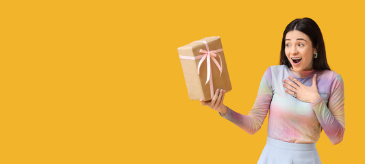 Surprised young woman with gift box on yellow background