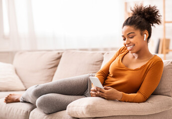 Obraz premium Weekend. African American Girl Using Phone Listening To Music In Earbuds Relaxing Sitting On Sofa Indoor. Selective Focus