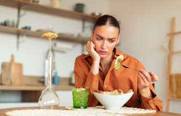 Sad young caucasian lady looking at her caesar salad and dont want to eat, sitting at kitchen...