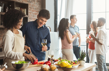 Home party. Multiethnic couple preparing dinner and drinking wine with friends in kitchen