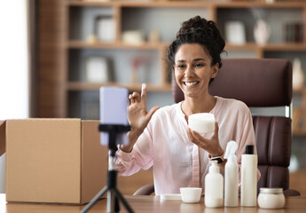 Smiling attractive young woman shooting video on smartphone, influencer blogger recording beauty vlog for social network, holding demonstrating cosmetic products, makeup haul, tutorial, home interior