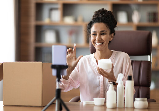 Smiling attractive young woman shooting video on smartphone, influencer blogger recording beauty vlog for social network, holding demonstrating cosmetic products, makeup haul, tutorial, home interior