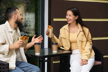 Young happy couple with tasty pretzels and cups of coffee sitting in cafe