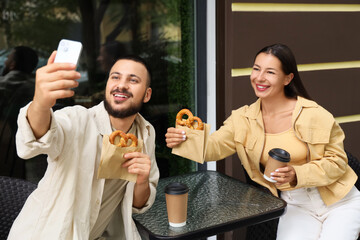 Young happy couple with tasty pretzels, cups of coffee and mobile phone taking selfie in cafe