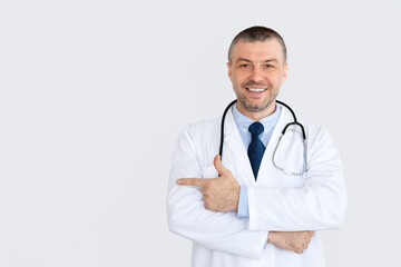 Fototapeta premium Useful Information. Portrait of smiling confident mature general practitioner pointing away at copy space standing isolated over white studio background, looking at camera. Doctor wearing stethoscope