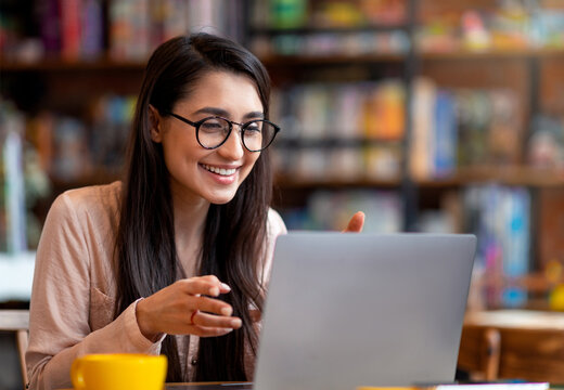 Positive arab woman having video conference on laptop, sitting at city cafe, talking and gesturing to computer webcamera. Online tutoring and remote distant communication concept - Powered by Adobe