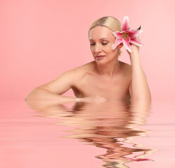 Beautiful woman with perfect skin and lily flower against pink background. Her reflection in...